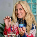Woman holding a bottle of 'dr.emil' multivitamin supplement and a glass of water indoors.
