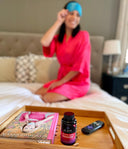 Woman in pink robe sitting on a bed with a tray of a book, bottle, and remote control.