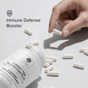 Hand picking up a supplement capsule with 'Immune Defense Booster' text and product name on a light background.
