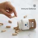 Hand picking up a supplement capsule from an open bottle labeled 'Immune Defense' on a marble surface.